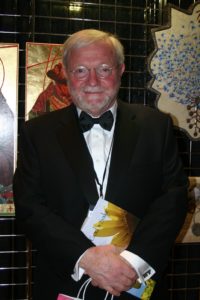 An older gentleman, with wire frame glasses and dressed in a tuxedo, stand facing the camera with his hands folded in front of him.