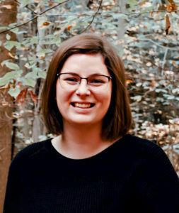 Tesia is a white woman with shoulder length brown hair wearing glasses. She is pictured smiling, wearing a black sweater, with leaves and trees in the background.
