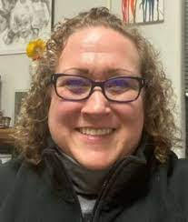 A White woman with dark blonde, curly shoulder-length hair wearing glasses smiles at the camera.