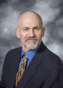 Smiling bald man with a grey goatee wearing a black suit jacket, blue shirt and patterned brown tie.